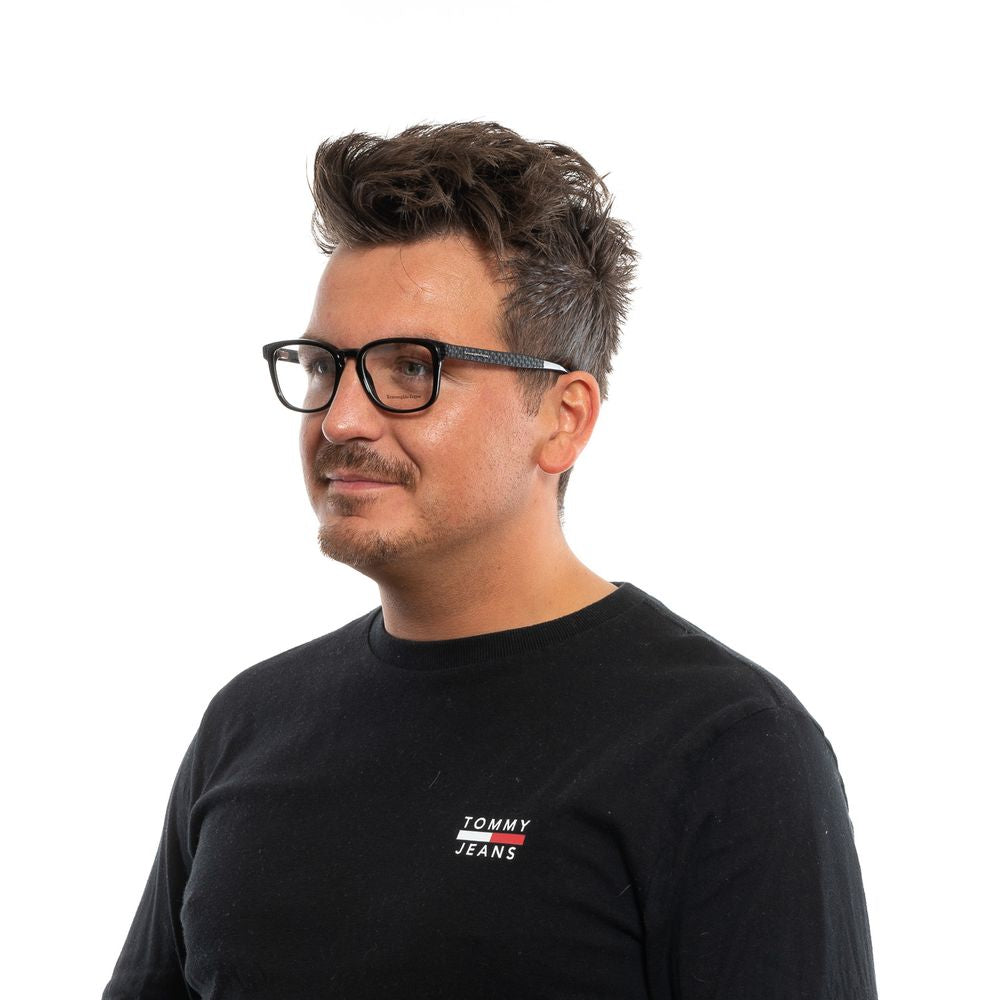 A man with short, dark, tousled hair and wearing Ermenegildo Zegna Black Plastic Glasses frames is dressed in a black shirt, facing slightly left and smiling gently against a plain white background.